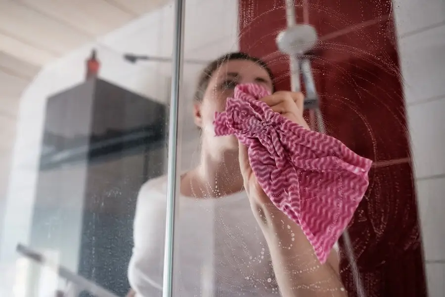 Woman cleaning a shower glass door with a pink cloth to remove water spots and soap scum.