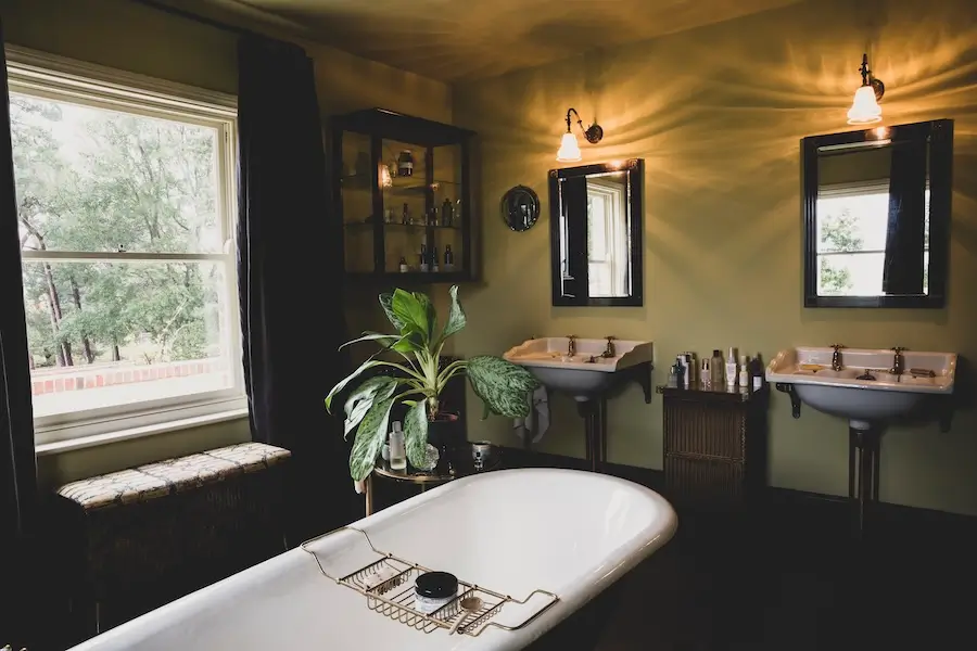 Vintage-style bathroom with a freestanding white bathtub, dual pedestal sinks with mirrors, green walls, and a large window letting in natural light.